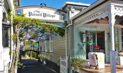 Discover the iconic entrance to Parnell Village in Auckland City, featuring a grand decorative arch, charming white heritage buildings, and lush leafy walkways.