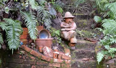 Explore a lush moss-filled garden on your Coromandel tour, featuring a whimsical child statue in a hat, ferns, pottery, and brick steps.