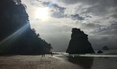 Golden sun rays shine through dramatic clouds above Coromandel beach, highlighting spectacular rock formations along the scenic shore.