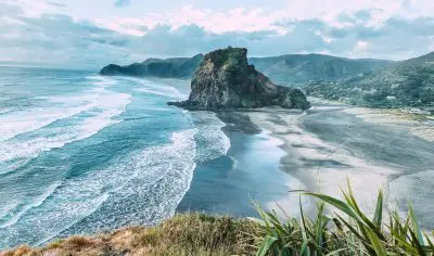 Stunning West Coast black sand beach, dramatic rocky outcrop, towering mountains, cloudy skies, and lush grass in the foreground.