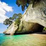 Cathedral Cove’s sandy beach, crystal-clear turquoise waters, iconic rocky arch and lush trees under a vibrant blue sky in New Zealand.