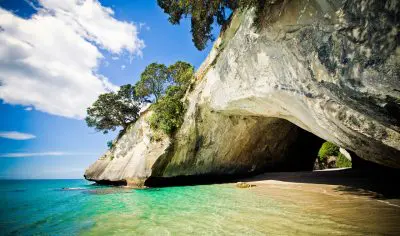 Cathedral Cove’s sandy beach, crystal-clear turquoise waters, iconic rocky arch and lush trees under a vibrant blue sky in New Zealand.