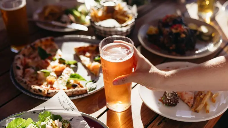 Hand holding a frosty beer on rustic wooden table, surrounded by gourmet pizza, fresh salads, and Margaret River adventure ambience.