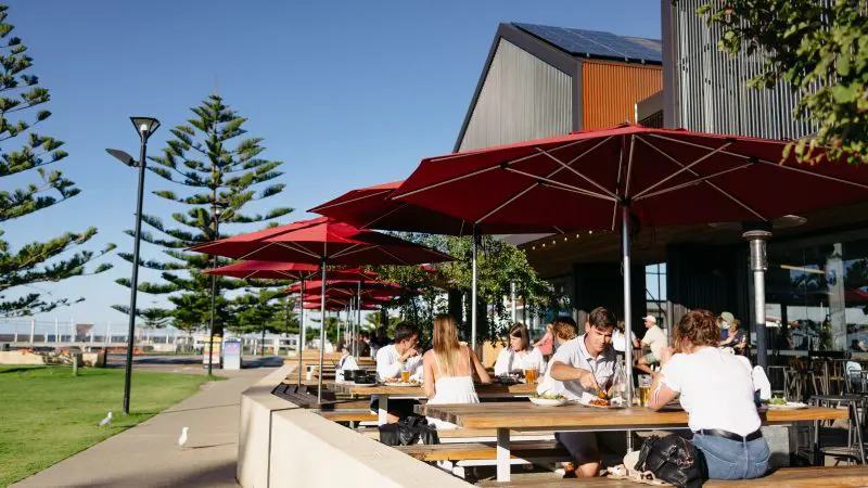 Visitors savouring gourmet meals outdoors beneath vibrant red umbrellas by a modern building on a Margaret River Wildlife Adventure.