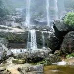 Spectacular tall waterfall pours over rugged cliffs in the Blue Mountains, surrounded by lush greenery on a sunset wilderness day tour.