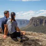 A couple enjoys panoramic mountain views from a rocky cliff on a 1 Day Blue Mountains Sunset Wilderness Tour adventure.