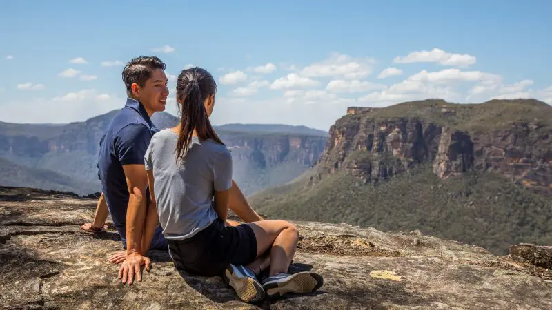 A couple enjoys panoramic mountain views from a rocky cliff on a 1 Day Blue Mountains Sunset Wilderness Tour adventure.