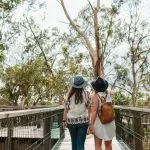 Two women in hats stroll together on a wooden boardwalk at Phillip Island Penguin Parade, lush tall trees surrounding the famous site.
