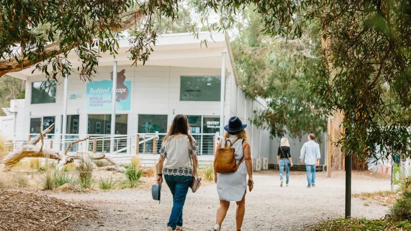 Two women approach a modern building, experiencing the ultimate Phillip Island Penguin Parade and Koalas Tour with exclusive viewing.