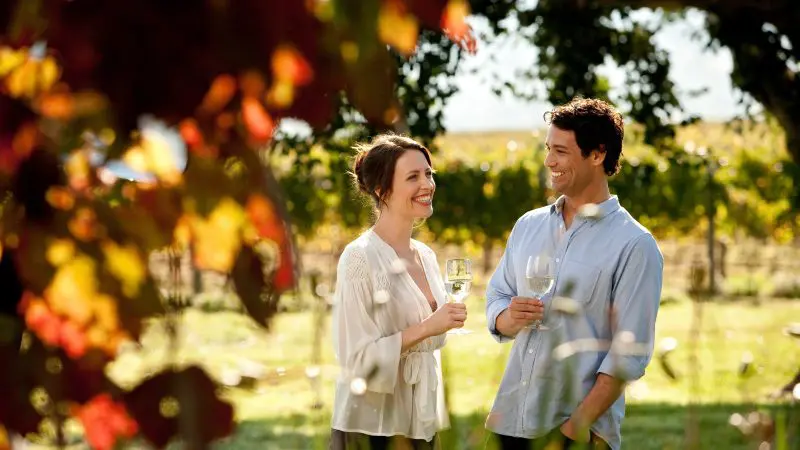Happy couple savouring white wine amid lush Hunter Valley vineyards on a sunny 1 Day Scenic Wine Tour, perfect for wine lovers.