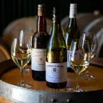 Three premium wine bottles and four elegant white wine glasses displayed on a rustic barrel, ideal for Hunter Valley wine tours.