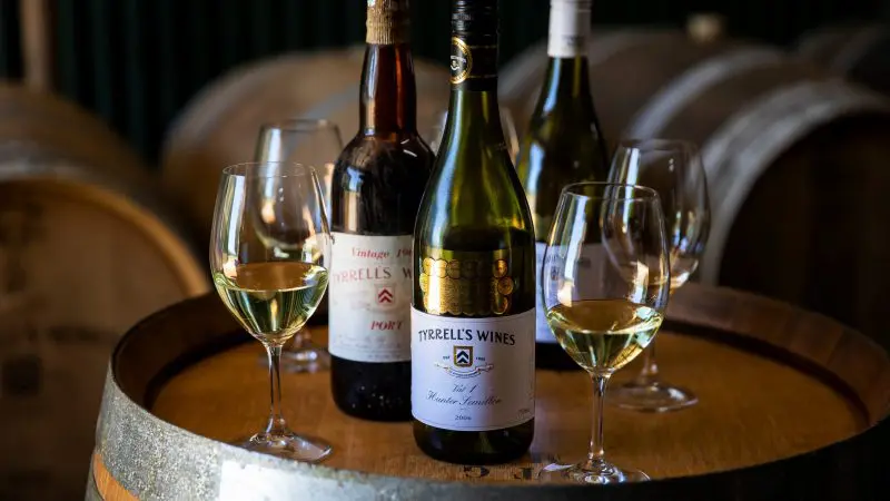 Three premium wine bottles and four elegant white wine glasses displayed on a rustic barrel, ideal for Hunter Valley wine tours.