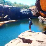 Adventurer with rucksack stands on rocky ledge during Kakadu Koolpin Tour, gazing over a pristine blue river and lush natural landscape.
