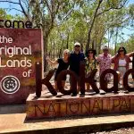 Group of six tourists smiling at the Kakadu National Park sign, eager to discover top Kakadu Tour Highlights amid lush greenery.