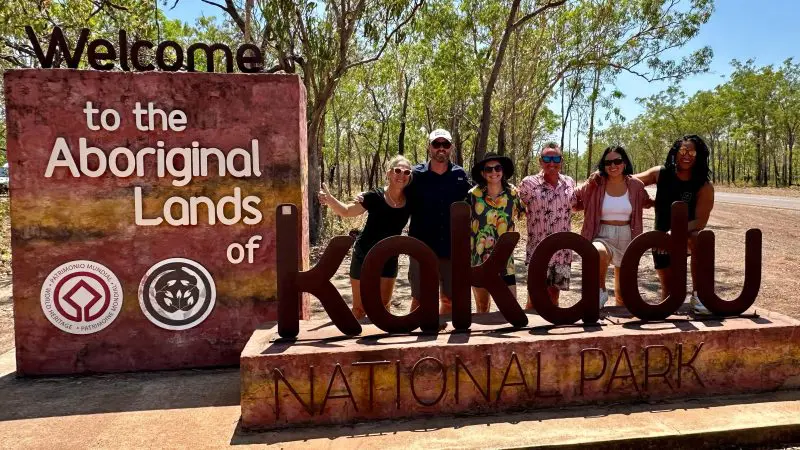 Group of six tourists smiling at the Kakadu National Park sign, eager to discover top Kakadu Tour Highlights amid lush greenery.