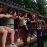 Happy group leans from a Puffing Billy Steam Train carriage, posing with friendly guard on the platform for memorable tour photo.