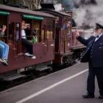 Train guard waving to excited children on Puffing Billy Steam Train, holding brass horn, with clouds of steam billowing in the background.