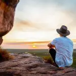 Traveller wearing a hat sits on a rugged rocky ledge, admiring Kakadu's breathtaking sunset over vast, dramatic cloudy landscapes.