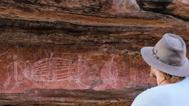 Visitor in a wide-brimmed hat studies remarkable ancient white Aboriginal rock art on vivid red stone, a top Kakadu Tour highlight.