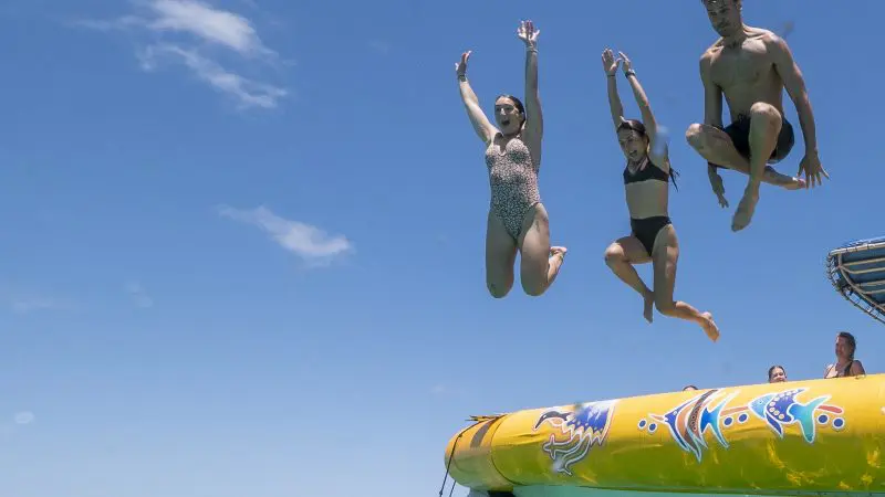 Three adventurers in swimming costumes leap from a yellow inflatable boat into the crystal-clear Great Barrier Reef, basking in sunny skies.
