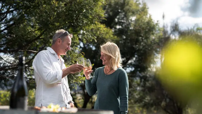 A smiling couple toasts with wine glasses aboard a Tasman Safaris Port Arthur Harbour Cruise, enjoying a scenic outdoor lunch.