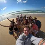 Friends pose for a bright selfie on an East Coast Explorer tour, smiling with rolling ocean waves and sunny skies in the background.