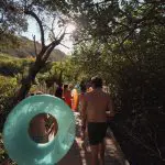 Group of holidaymakers in swimming costumes stroll along a sunlit promenade, carrying lilos on their East Coast Explorer beach holiday.