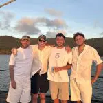Four men in white outfits enjoy drinks and smile on a boat tour, East Coast Explorer, with lush green hills and sparkling water behind them.