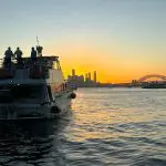 Sydney Harbour sunset cruise with people on a boat, golden sky reflecting on water, iconic Sydney skyline illuminated in the background.