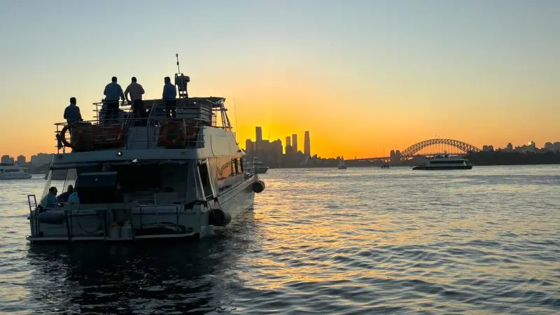 Sydney Harbour sunset cruise with people on a boat, golden sky reflecting on water, iconic Sydney skyline illuminated in the background.