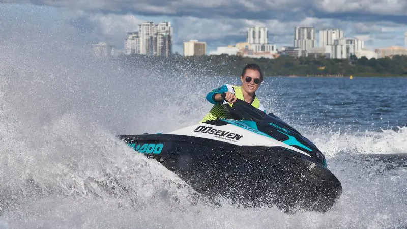 Jet ski rider speeding by Thunderball Shipwreck, sparkling blue waters, city skyline, lush trees; thrilling Bahamas adventure scene.