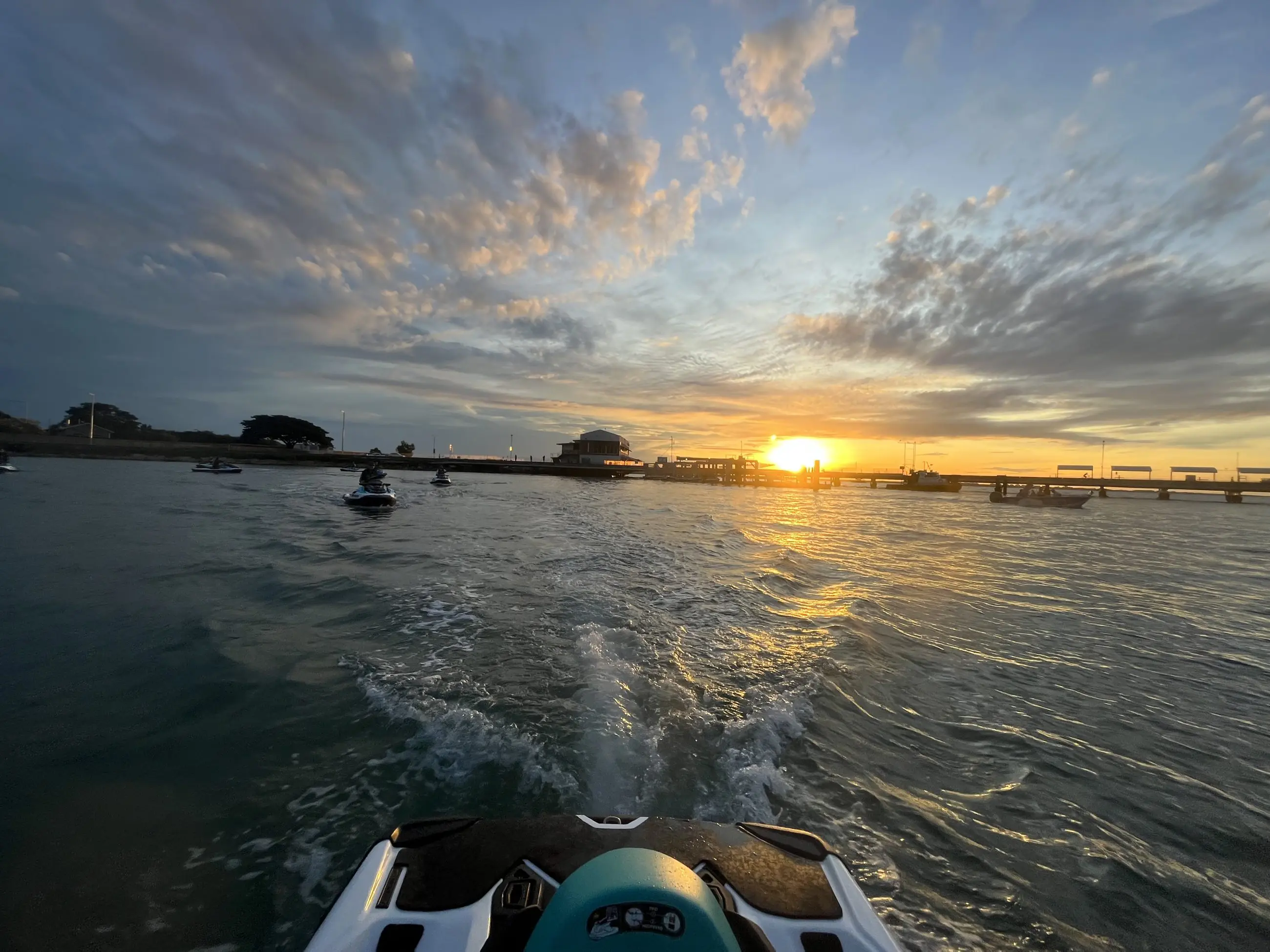 A jet ski speeds across the water towards a pier at Honey Rider Sunrise, beneath dramatic clouds and a glowing sun on the horizon.