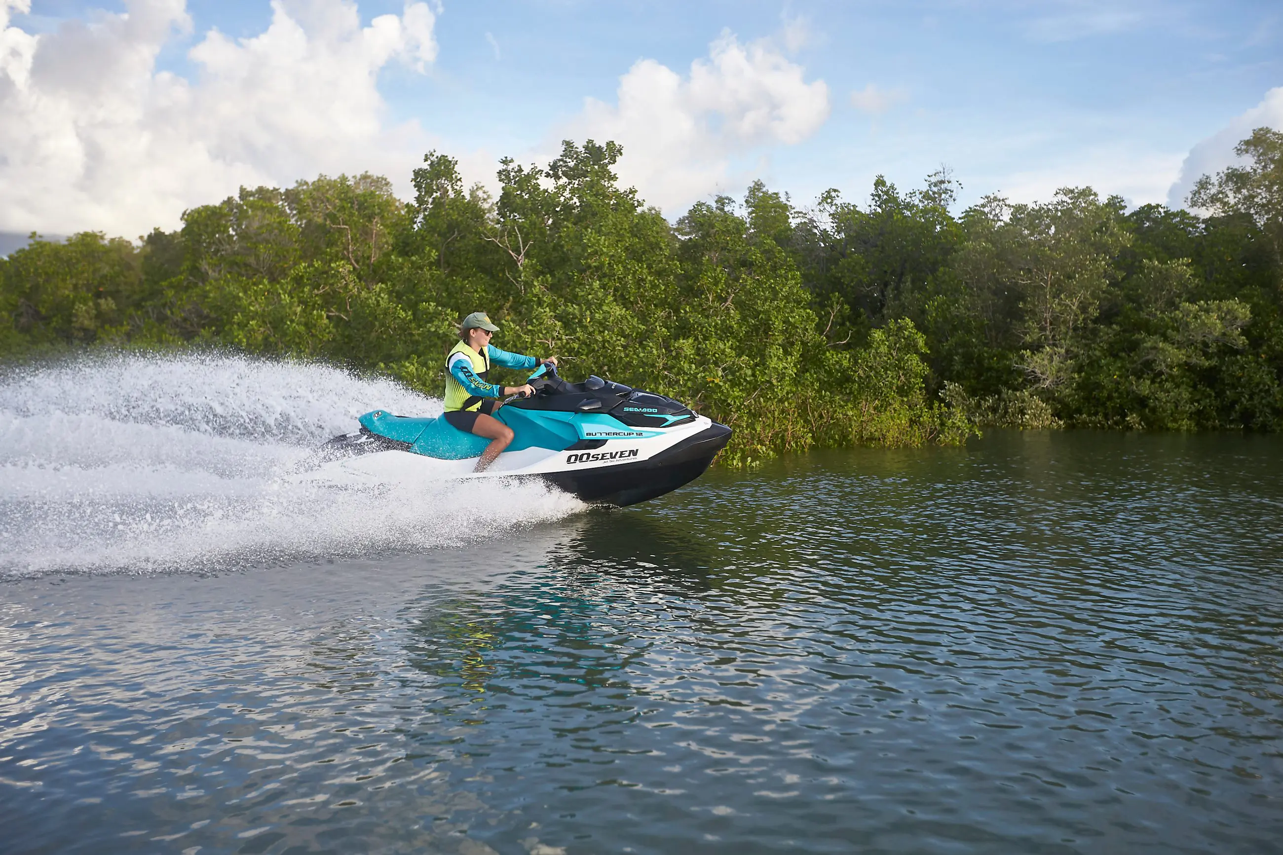 Person speeding on a blue jet ski, Casino Royale style, on a river bordered by lush green trees, wearing life jacket and cap.