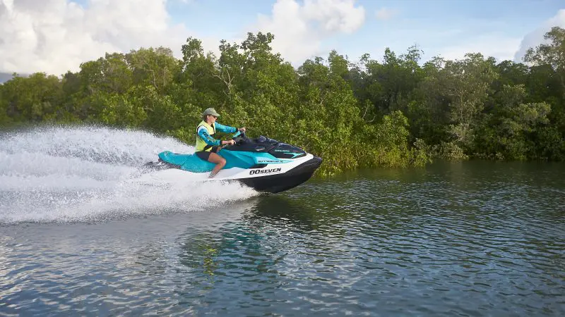 Person speeding on a blue jet ski, Casino Royale style, on a river bordered by lush green trees, wearing life jacket and cap.