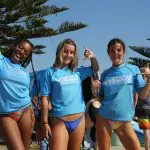 Three women in blue rash vests smile by surfboards, set for an epic 6 Day Surf Trip from Sydney to Brisbane, surfing Australia’s coast.