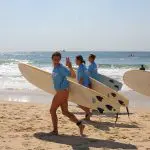 Group of four surfers in blue shirts with surfboards on a sunny Sydney to Brisbane trip; one smiles and waves at the camera.