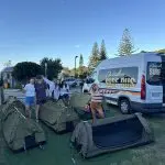 Four travellers by tents and the iconic Australian Surf Bus on a 6-Day Sydney to Brisbane Surf Trip beneath clear blue skies.