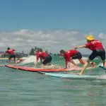 Four surfers in matching red shirts catch waves under a sunny sky on a 6-Day Sydney to Brisbane Surf Trip, rolling green hills behind.