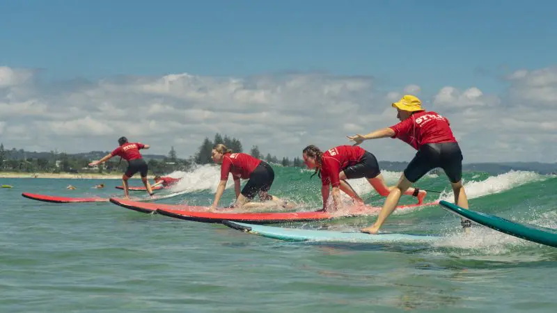 Four surfers in matching red shirts catch waves under a sunny sky on a 6-Day Sydney to Brisbane Surf Trip, rolling green hills behind.