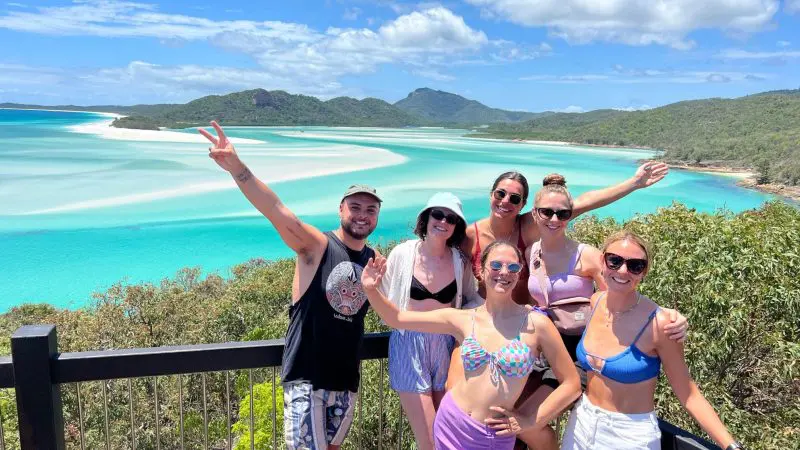 Group of five friends smiling on a Summer Jo Day Charter overlook, vibrant turquoise water and lush green hills in the background.