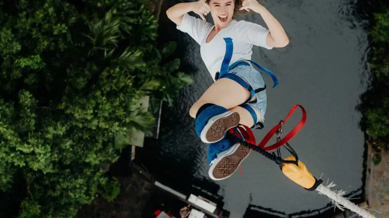 Smiling woman gives thumbs-up mid-air while bungee jumping over blue water at Australia’s Only Bungee Skypark, aerial view.