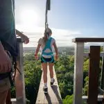 Adventurer in safety harness readies for thrilling zip wire ride at Walk The Plank Skypark, above vibrant green landscape on sunny day.