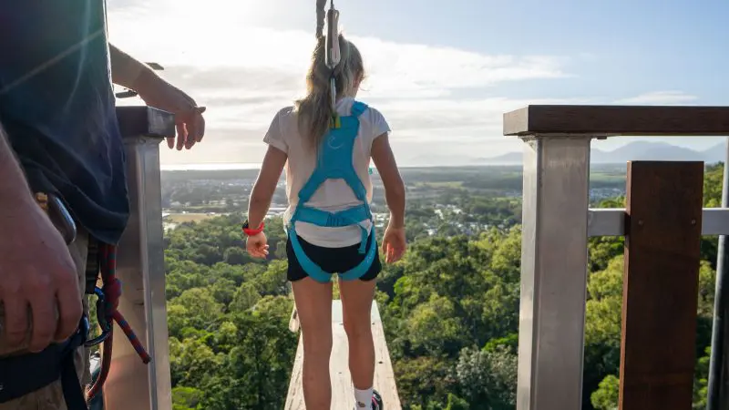 Adventurer in safety harness readies for thrilling zip wire ride at Walk The Plank Skypark, above vibrant green landscape on sunny day.