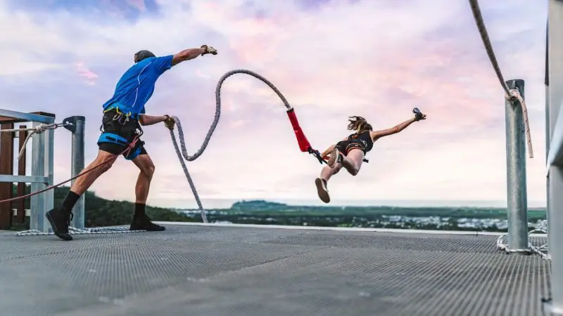 Thrill-seeker starts bungee jump at Roof Jump Skypark, instructor secures safety cord, dramatic sunset in the background.
