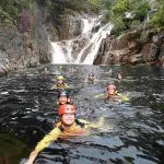 Adventurers in helmets swim through a rocky river beside a stunning waterfall on the Behana Gorge Canyoning Tour in Queensland.