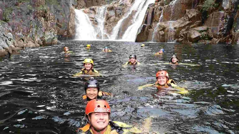 Adventurers in helmets swim through a rocky river beside a stunning waterfall on the Behana Gorge Canyoning Tour in Queensland.
