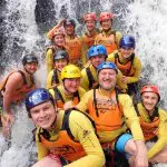 Smiling adventurers in helmets and life jackets pose by picturesque Crystal Cascades waterfall during an unforgettable canyoneering tour.