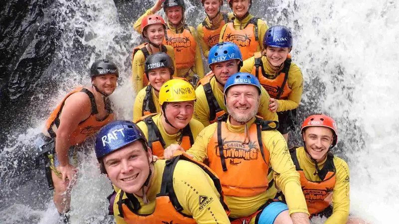 Smiling adventurers in helmets and life jackets pose by picturesque Crystal Cascades waterfall during an unforgettable canyoneering tour.