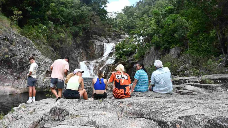 Visitors unwind on sunlit rocks beside a stunning waterfall, enveloped by lush rainforest on the Creek to Coral Mission Beach Day Tour.