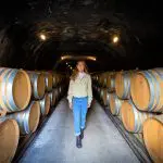 A woman explores a dimly lit wine cellar tunnel, walking between stacked barrels on a luxurious gourmet wine tasting tour experience.